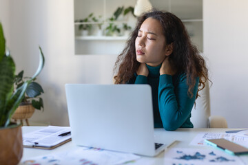 Feeling exhausted. Frustrated Asian woman looking exhausted and massaging her neck while sitting at her working place