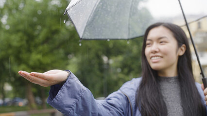 Smiling asian woman feeling rain with hand, enjoying fresh weather, city walk