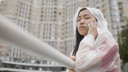 Upset asian woman in raincoat talking on phone in rainy city, calling taxi