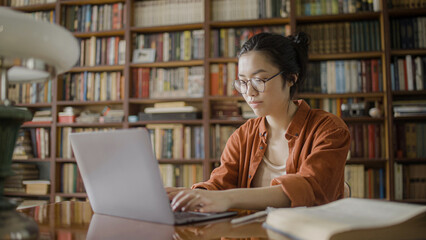 Focused young woman student spending time in university library, doing research, typing on laptop