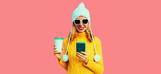 Winter portrait of happy smiling young woman with smartphone and coffee cup wearing yellow knitted sweater, white hat with pom pom, heart shaped sunglasses on pink background