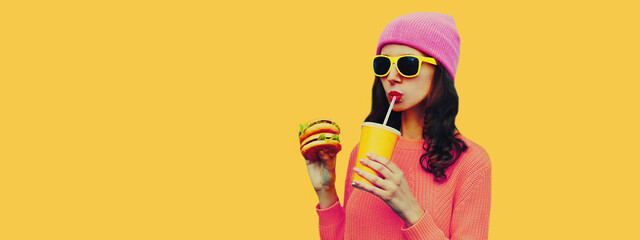 Portrait of stylish young woman drinking juice with burger fast food on yellow colorful background