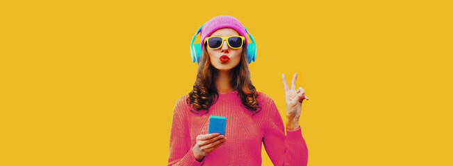 Portrait of stylish young woman in headphones listening to music with smartphone and blowing her...