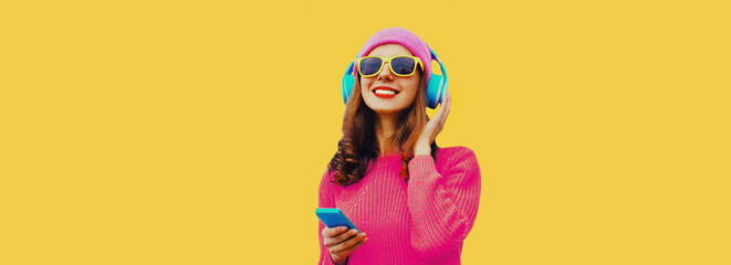 Portrait of happy smiling modern young woman in wireless headphones listening to music with smartphone wearing knitted sweater, pink hat on yellow background - obrazy, fototapety, plakaty