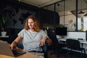 Young man using laptop and earphones while sitting by table in office