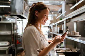 Young blonde woman using cellphone while working in restaurant kitchen
