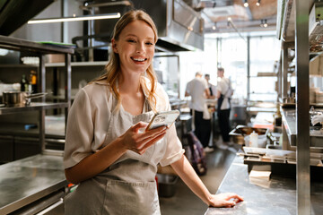 Young blonde woman using cellphone while working in restaurant kitchen - 559723803