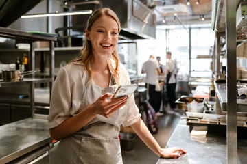 Tuinposter Young blonde woman using cellphone while working in restaurant kitchen © Drobot Dean