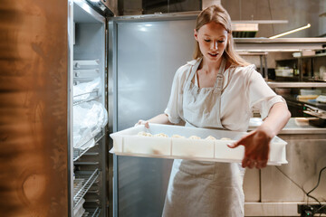 Young blonde woman putting food in refrigerator working in kitchen