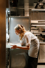 Young blonde woman putting food in refrigerator working in kitchen - 559723839