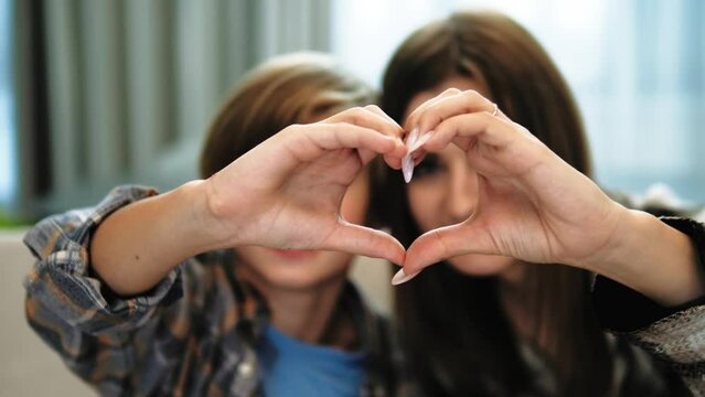 Close up portrait happy sincere  family mother and teenager son boy making heart gesture with fingers, showing love or demonstrating sincere feelings together indoors, looking at camera
