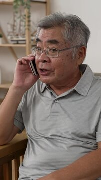 Vertical Screen: shocked old asian man sitting on sofa and answer smartphone call while hearing bad news. confused grandpa in glasses worried about his family