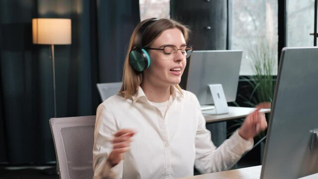 Amazed happy businesswoman female office worker in wireless cool headphones dance at office place emotional young girl feel euphoric have fun, businesswoman sitting at desk working on computer pc.