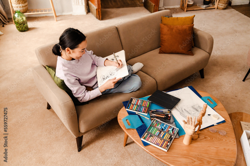 Wall mural top view of cheerful asian woman artist drawing in sketchbook while sitting on couch at home