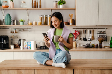 Smiling asian woman using mobile phone and drinking coffee while sitting in kitchen