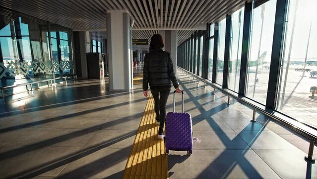 Girl with suitcase walks at airport along corridor with large windows. Back of young business woman passenger walking down walkway. Fly in airplane. Go to rest travel. Vacation and trip, tourism.