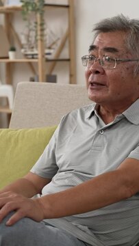 Vertical Screen: Emotional senior man watching tv sitting on couch with remote controller at home. mad old male with confused face and talking himself on sofa in apartment