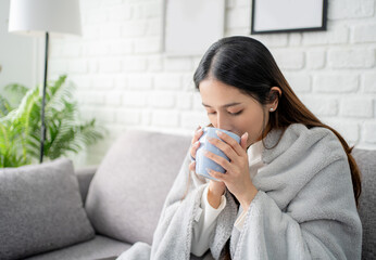 Cold young woman enjoying a mug of hot winter coffee as she wrapped up in blanket in a warm on her...
