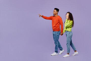 Full body side view young couple two friend family man woman of African American ethnicity in casual clothes hold hands walk go together point finger aside isolated on pastel plain purple background