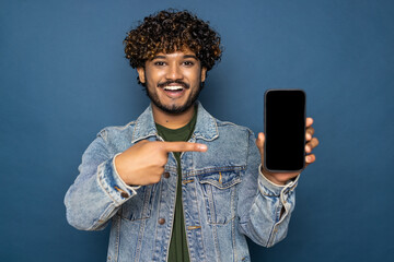 Happy excited indian or arabian guy, in casual wear, holds smartphone with blank white screen for...