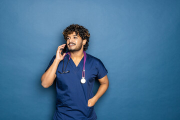 Smiling indian male doctor in white coat with stethoscope calling on smartphone over blue background
