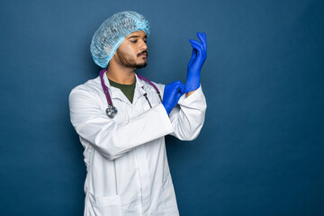 Indian doctor or physician male looking at hand with focused expression as putting on blue surgical latex glove isolated on blue background