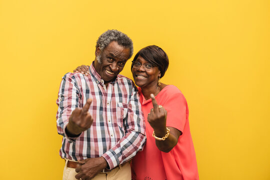 Smiling senior couple showing obscene gesture against yellow background
