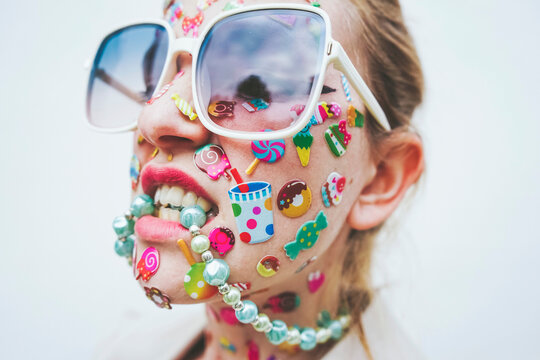 Woman with multi colored stickers on face biting necklace in front of white background