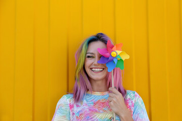 Happy woman wearing tie dye t-shirt holding colorful pinwheel toy in front of yellow wall