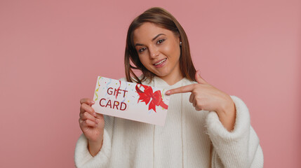 Young woman in sweater showing pointing on gift discount certificate voucher coupon for store holidays sale. Pretty girl with shopping christmas surprise card isolated alone on pink studio background