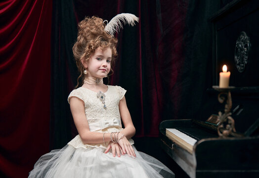 Portrait of cute little girl, child in image of medieval royal person, princess in fabulous dress sitting at the piano. Concept of historical remake, comparison of eras