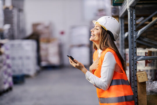 A warehouse worker is using cellphone and laughing on message while standing in facility.