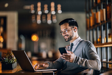 A happy young elegant entrepreneur is sitting in a restaurant and typing report while using phone.
