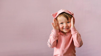 Portrait of toddler girl in jacket with cat ears on pink background.