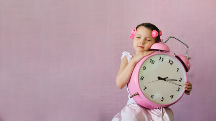 Funny emotional kid girl with curlers on her hair and big pink alarm clock. Copy space. Closed eyes.