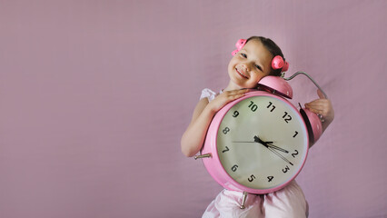 Funny emotional kid girl with curlers on her hair and big pink alarm clock. Copy space.