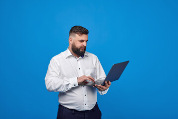 Naklejka na ściany i meble A young man on a blue background holds a laptop in his hands. A bearded man works at a laptop on a blue background.