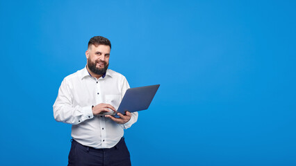 Obraz na płótnie Canvas A bearded man holds a laptop in his hands on a blue background. A solid man in a white shirt works at a laptop and looks into the camera.