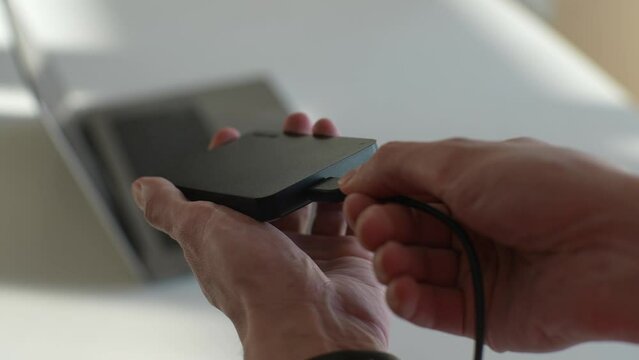 Close-up cropped shot of unrecognizable man connecting USB cable to black external hard drive on background of laptop on desk. Connect home storage to computer. Shooting in slow motion.