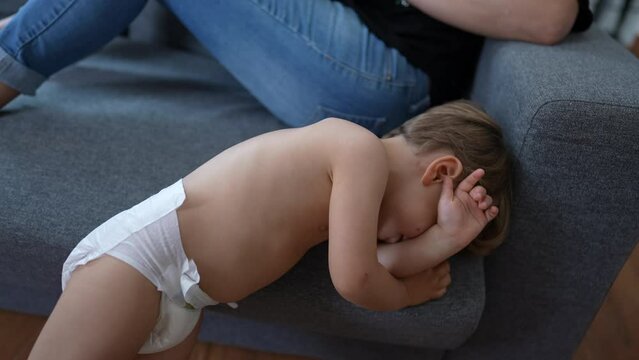 One tired little baby toddler wearing diapers rubbing face on couch