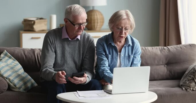 Couple of retirees managing family budget, paying bills and taxes using on-line electronic bank website on laptop, analyzing expenses sit on sofa at home. Finances control, pension, monthly payments