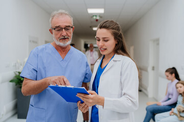 Older doctor giving advise to his younger colleague, discussing at hospital corridor. Health care...