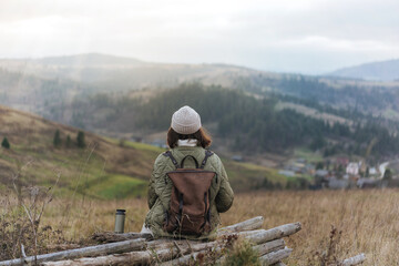 View from the back of hiker girl wearing backpack and beanie hat sitting on edge of cliff and enjoying  mountains on the sunset