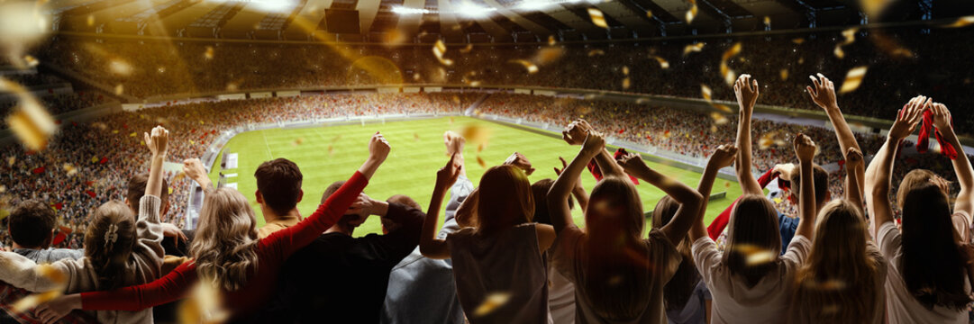 Back view of football, soccer fans cheering their team with flag and posotove emotions at crowded stadium at evening time. Concept of sport, hobby, leisure time, football