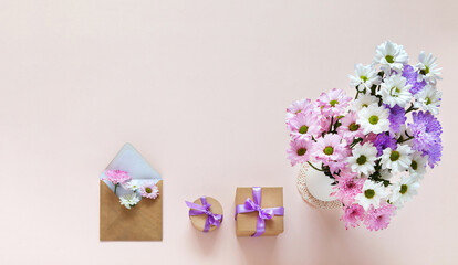 Festive greetings with bouquet of delicate chrysanthemums, gifts in holiday boxes and envelope with wishes on  light pink background. Celebration of Valentine's Day, March 8, anniversary or birthday
