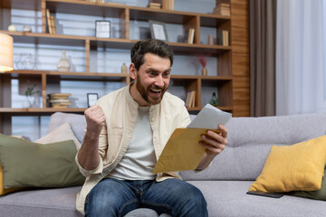 Joyful man at home holding envelope with notification message reading and smiling happy with news sitting on sofa inside living room.