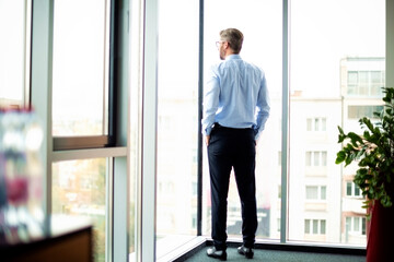Shot of thinking businessman standing at the office and looking out of city