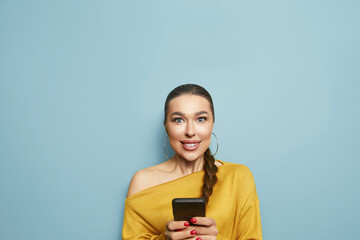 Smiling young brunette woman girl in yellow sweater posing isolated on pastel blue wall background studio portrait. People lifestyle concept. copy space. Using mobile phone, typing sms message