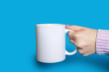 Obraz premium A hand holds a white cup with a hot drink on a blue background. The hand of a woman dressed in a sweater holds a large ceramic cup against a bright blue background. Concept of drinking hot drinks
