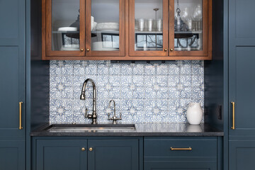 A kitchen sink with a beautiful pattern tiled backsplash with a chrome faucet, black granite countertops, and surrounded by blue and wood cabinets.	